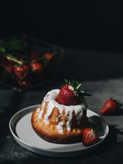 Cake with white cream and fresh strawberries, lying on a white plate on a dark background. The sun is shining on the dish. In the background, a basket of berries. Strawberry dessert.