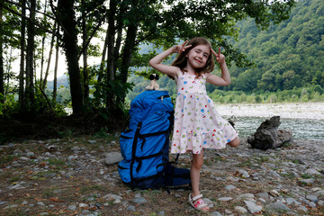 A large backpack for an adult stands next to a little girl against the backdrop of trees and a river, on a sunny day.