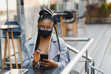 young pretty african american women in a medical protective mask outside in cafe. african girl with tablet and phone.