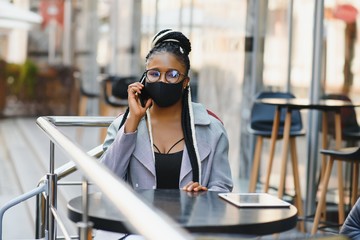 Fototapeta premium young pretty african american women in a medical protective mask outside in cafe. african girl with tablet and phone.