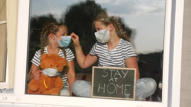 stay at home coronavirus. kids in medical mask in home with a toy teddy bear. kids holding a sign with the inscription stay at home. quarantine sitting by the window covid 19. coronavirus concept