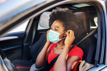 Young attractive african woman sitting in her car and putting mask on her face. Covid-19 outbreak...
