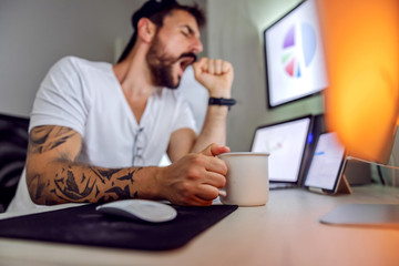 Young tired bearded freelancer holding mug with coffee and yawning while sitting at home office. Selective focus on hand.