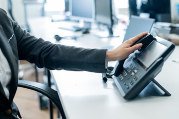 Closeup female hand on landline phone in office. Faceless woman in a suit works as a receptionist answering the phone to customer calls.
