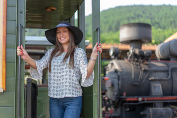 Beautiful brunet woman wearing black hat, boarding in train. Posing for camera. Old locomotive in background. 