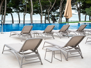 Empty sunbeds near empty swimming pool with conifers on background.