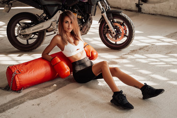 Fototapeta na wymiar Pretty and attractive sportswoman posing in a light studio sitting on the floor with a large orange punching bag. She is wearing a black and white tracksuit and she has orange boxing gloves.