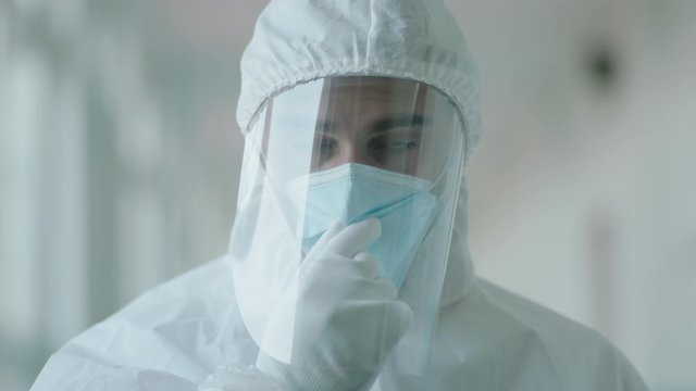 This stock video shows a close-up of a male doctor or medical researcher wearing protective gear, mask and face shield in a laboratory and looking at the camera
