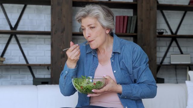Elderly woman eats her vegetable salad sitting on the sofa at home