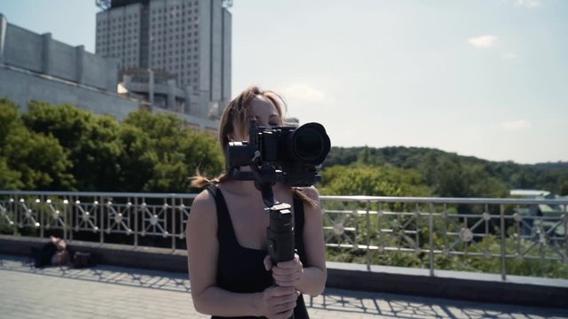 Videographer shoots on camera with hand held tripod. Action. Female photographer takes pictures with professional camera with manual stabilizer. Shooting video on roof with view of city