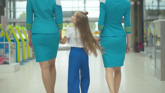 Back view of happy Caucasian girl holding hands of professional stewardesses and walking to airport exit. Brunette child strolling with flight attendants after arrival. Chaperoning concept.