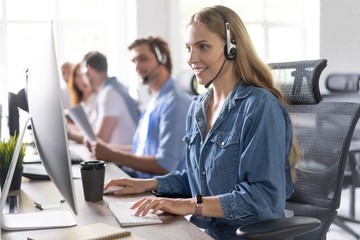How can I help you? Beautiful call center workers in headphones are working at modern office. - obrazy, fototapety, plakaty