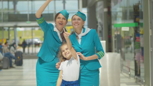 Charming little girl standing with stewardesses next to airport and looking at plane takeoff. Portrait of happy Caucasian child enjoying sunny day with professional flight attendants before departure.