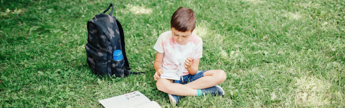 Elementary school boy sitting in park outdoors doing school homework. Child kid reading book outside. Self education learning studying. Early development for children. Web banner header.