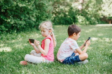 Boy and girl play games on smartphones outdoors. Kids digital gadget screen addiction. Children friends playing online sitting on grass in park. Contemporary problem of loneliness together.