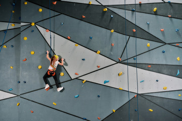Girl in safety equipment and harness training on the artificial climbing wall indoors. Bouldering...