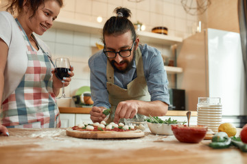 Passionate about pizza. Young couple making pizza together at home. Man in apron, professional cook adding basil on the dough while woman looking at him, drinking wine. Hobby, lifestyle