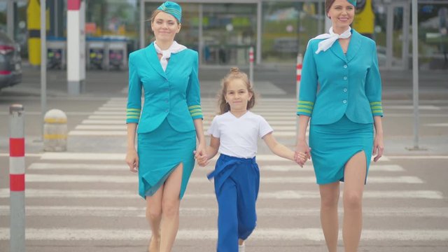 Wide shot portrait of happy Caucasian child walking with stewardesses out of airport and smiling. Cute little girl strolling with slim confident woman in flight attendant uniform outdoors and leaving.