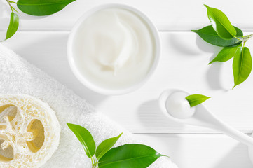 Natural herbal cosmetic cream, loofah sponge and towel on white wooden background with green leaves. Organic cosmetics concept.