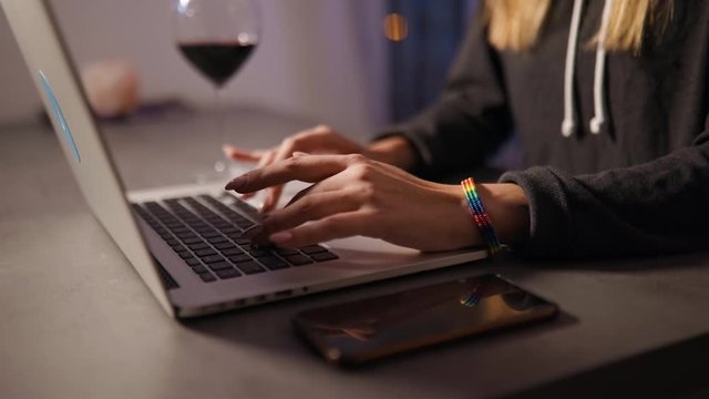 Close view of female hands typing on laptop wearing rainbw LGBT bracelet with glass of red wine and mobile phone on table