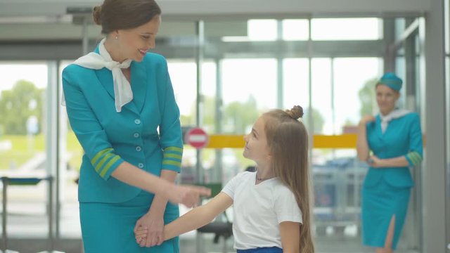 Charming cheerful woman in stewardess uniform talking to cute little Caucasian girl in airport. Portrait of beautiful flight attendant accompanying kid in international trip.