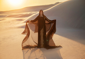 A mysterious woman in a black long dress stands in the desert. Luxurious clothes gold accessories...