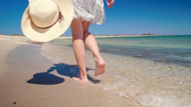 Female Foot Steps on Beach. Barefoot Woman Walks along Sea Coast with Hat in Summer Sunny Day. Holiday and Vacation on Chrysi, Crete, Greece. 4K Handheld Close up Tracking Backside Shot