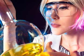 Portrait of a chemist girl. A blonde woman in safety glasses holds a flask of yellow reagent. Blogger-chemist. A woman demonstrates visual chemical reactions. Chemistry teacher.
