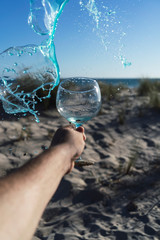 Mano sujetando una copa y arrojando bebida azul a a la playa