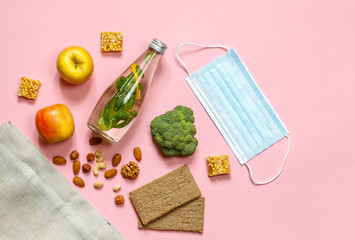 Fototapeta na wymiar Layout with a protective mask, a bottle of water, apples, broccoli and nuts laid out of the bag. pink background with space