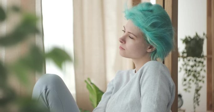 Woman with mint hair color enjoying fresh air and pleasant weather by sitting in front of open window