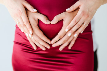 Concept of pregnancy and motherhood, unity of the family. Husband and wife together. A man holds the belly of his beloved pregnant woman. She puts her hands on top of his hands. Heart shaped fingers
