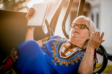 Elderly woman in blue dress is lying on the swing and using a digital tablet