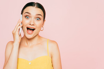 excited brunette young woman talking on smartphone isolated on pink