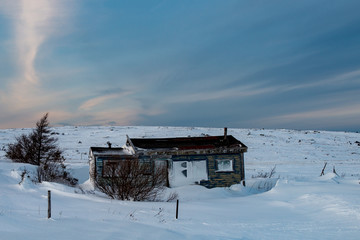 A small abandoned shack on the side of a country road. The old building is covered in snow. The meadow surrounding the architecture is also covered in heavy snowfall. The sky is blue with wispy clouds