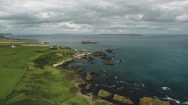 Rocky shore aerial view: green grass meadows and fields. Water surface stretches to horizon with grey clouds. Alone building on cliff. Northern Ireland panoramic landscape. Footage shot in 4K, UHD