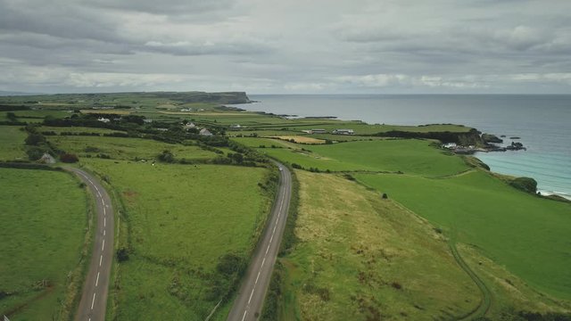 Aerial view road green meadows rise up drone shoot. Car drives along farmland way in Irish coutryside. Dramatic summer scenery of Northern Ireland in pastel tones. Footage shot in 4k, UHD