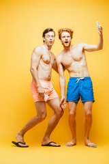 full length view of two shirtless men in shorts grimacing while looking at camera and taking selfie on smartphone on yellow