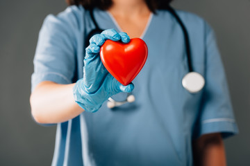 cropped view of doctor with stereoscope in latex glove holding red heart isolated on grey