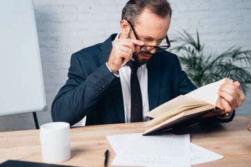 bearded lawyer in suit touching glasses while reading book in office