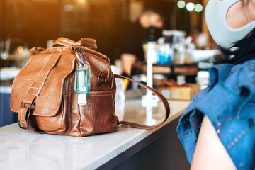Mini portable alcohol gel bottle to kill Corona Virus(Covid-19) hang on a brown leather shoulder bag on counter in coffee shop.New normal lifestyle. Health care concept. Selective focus on alcohol gel