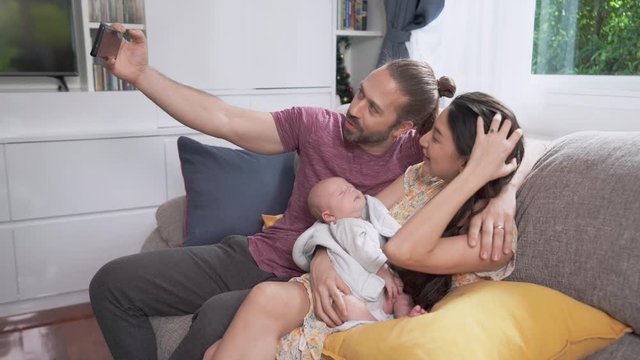 Smiling Caucasian Man using smartphone taking home video of his wife and sleeping newborn baby son. Father kissing mother and cute infant child boy with love. Happy Family and baby healthcare concept.