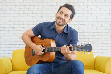 Happy man playing guitar while sitting on sofa in living room, Enjoying carefree time at home.