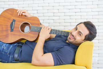 Happy man playing guitar while sitting on sofa in living room, Enjoying carefree time at home.