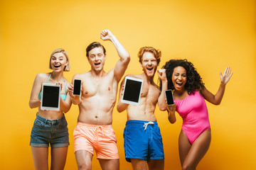 excited multiethnic friends in summer outfit showing digital devices with blank screen on yellow