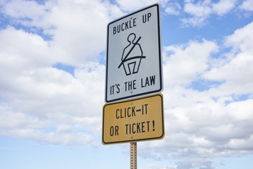 Buckle up sign and 'Click It or Ticket' warning sign against blue sky and clouds.