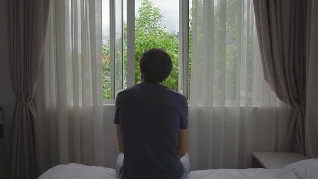 A young man in a mandatory quarantine. He sits and looks outside the window. He stays in one room for two weeks. He did not shave for long time, he is stressed and upset. Staying in quarantine concept