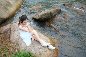 Young Asian girl sitting wearing white dress alone on the rock beside the stream in tropical rainforest.