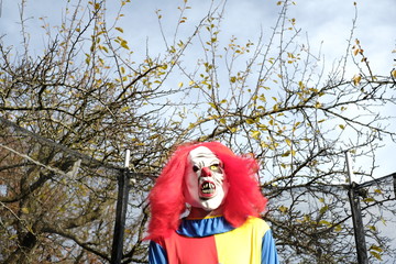  Halloween holiday. Evil clown  in a trampoline on autumn trees and blue sky background. Creepy clown costume. Autumn holidays in October. Festival and carnival concept. Horror and fear.