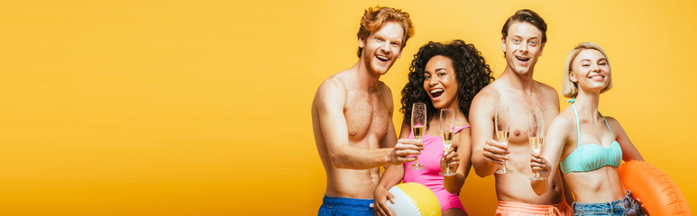 horizontal image of young multicultural friends holding champagne glasses, inflatable ring and ball while looking at camera isolated on yellow